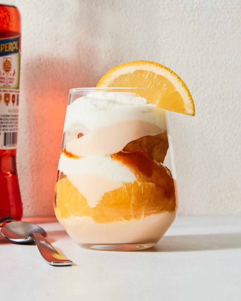 a stemless wine glass filled with vanilla cake, whipped cream, aperol spiked jam, and pudding with an orange slice on the rim