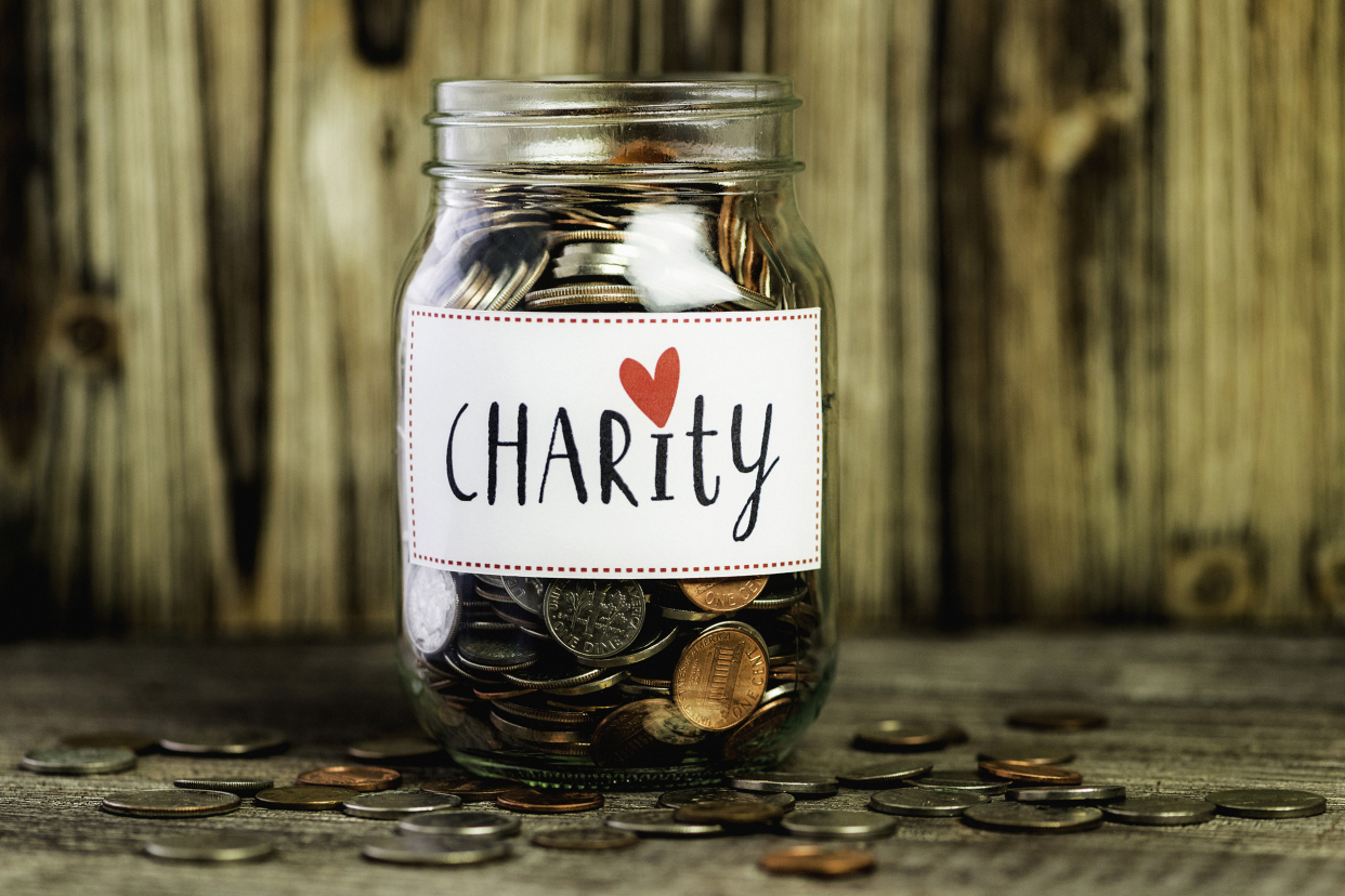 Opened glass mason jar with 'Charity' label sticker on it, filled with U.S. coins, surrounded by more coins on a wooden table with a wooden background