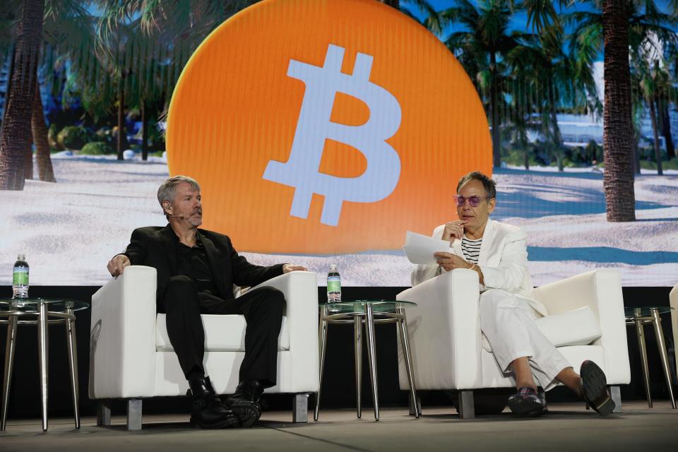 Michael saylor, dressed in all black, sits on a white couch, next to another man in all white, with a giant orange Bitcoin logo behind them.