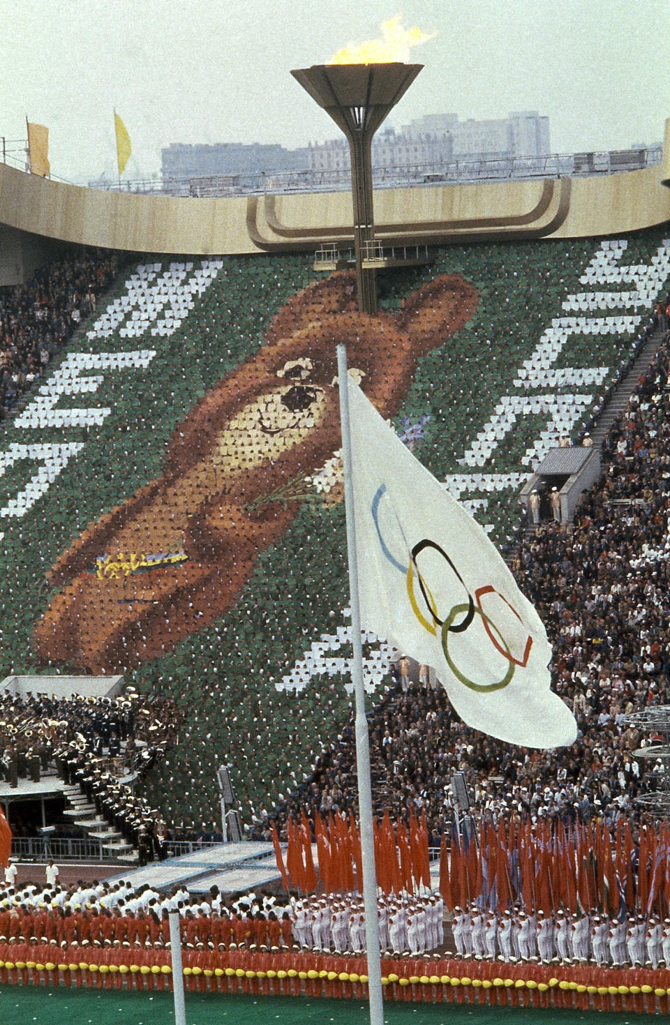 FILE - Members of the crowd hold up 3,500 cards to create an image of Misha the Bear Cup, the mascot of the Moscow Olympic Games at the Lenin Stadium in Russia on July 19, 1980. Above burns the Olympic flame. The 1980 Summer Olympics in Moscow brought much attention to the Soviet Union, for better and for worse. The Games did show that the USSR could be comfortable, high-spirited and welcoming to visitors despite laborious visa procedures. But dozens of countries, including the United States and sports powers Canada and West Germany, boycotted the event in protest of the invasion of Afghanistan. (AP Photo/File)