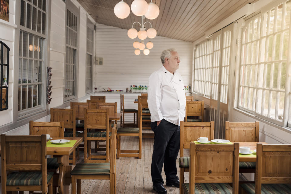 A man in a chef's uniform stands in an empty, rustic dining area with wooden chairs and tables, looking out the window