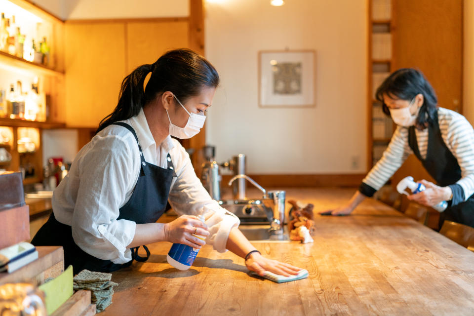 Two women wearing a mask, Carefully wipe a counter  and enthusiastically disinfect in preparation for opening.Cafe made of natural wood.