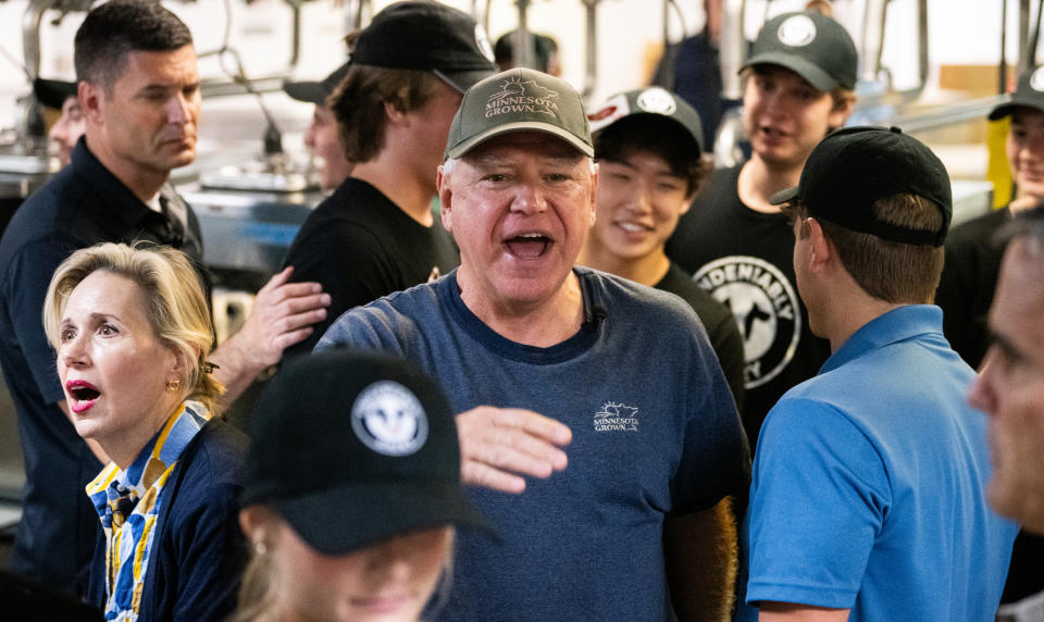 Several people, including Tim Walz, casually dressed in a blue t-shirt and hat, and interacting with others at a crowded event.