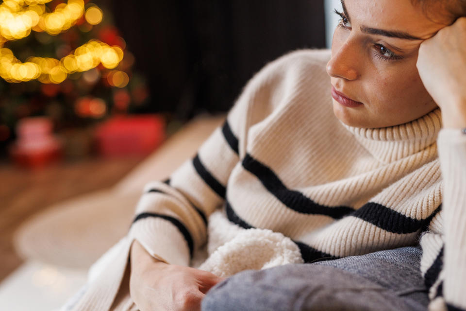 Portrait of anxious young woman sitting on the sofa, under a cozy, warm blanket, contemplating about spending Christmas holidays alone.