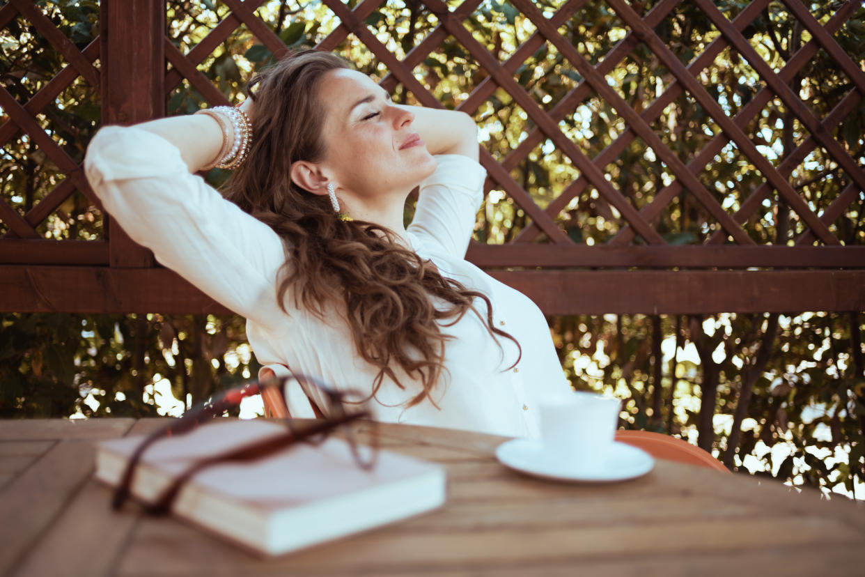 A woman smiles with eyes closed near reading glasses, a book and a coffee cup while leaning back into a chair with her hands clasped behind her head.