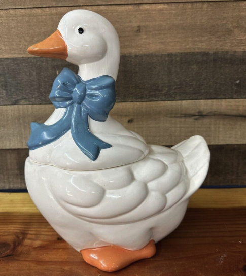 Ceramic goose-shaped container with a bow around its neck, sitting on a wooden surface