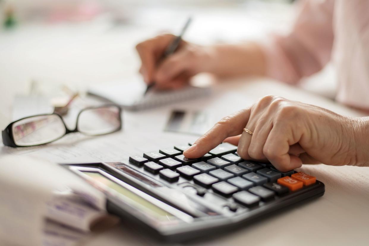 Senior woman's hand using a calculator with the other hand writing on a notepad with a blurred background of reading glasses, papers, and other items on a table