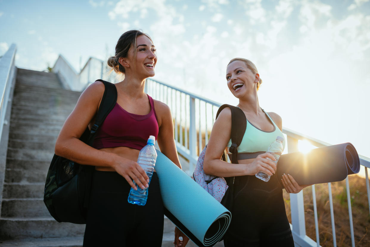 Two women holding their yoga mats and water bottles glow after finishing a class with stairs behind them.