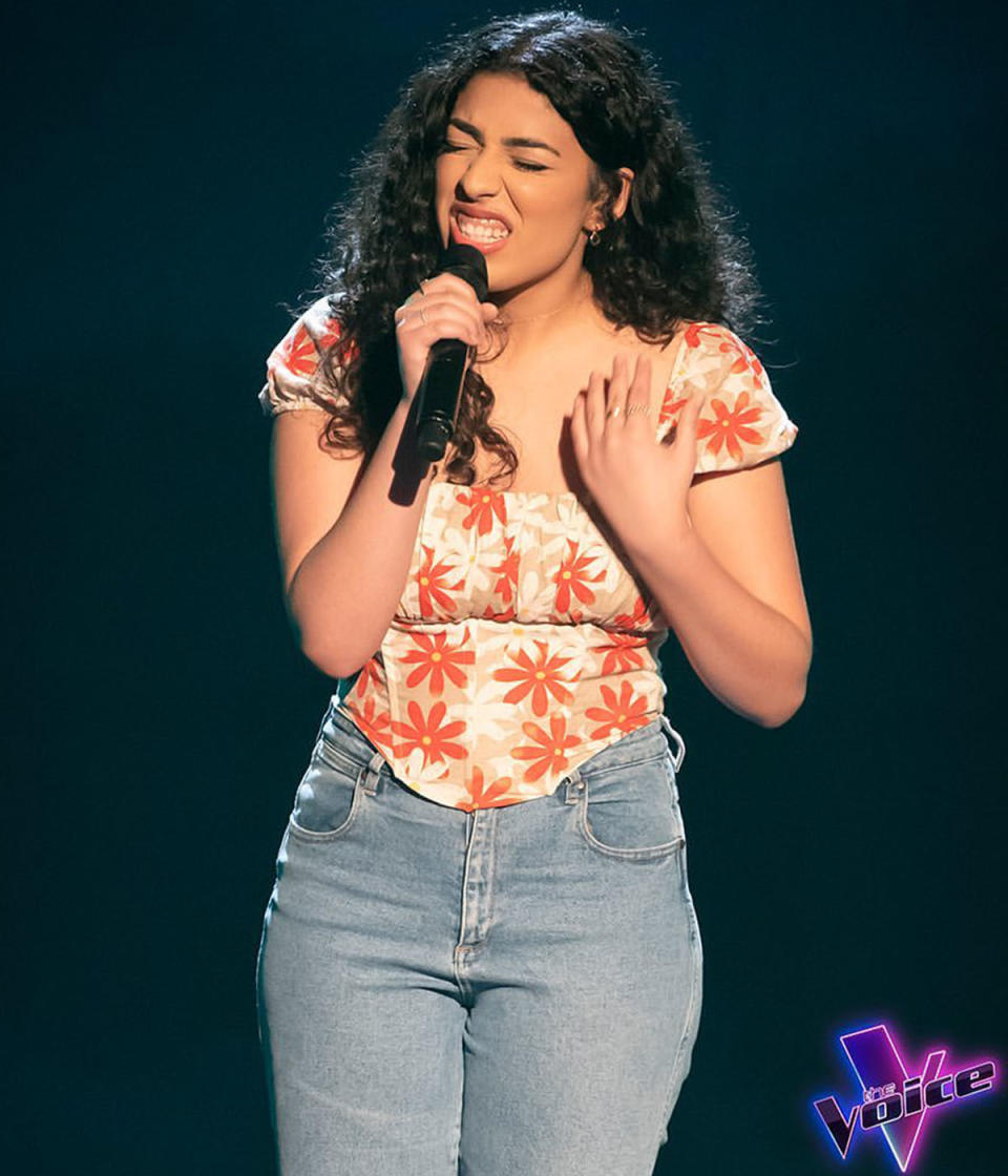 Alisha George, The Voice contestant, is pictured singing into a microphone. She wears jeans with an orange and white floral top.