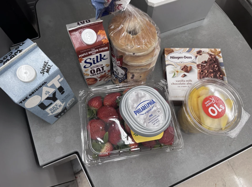 Groceries on a checkout counter, including oat milk, Silk maple brown sugar oat milk, bagels, Philadelphia cream cheese, strawberries, Häagen-Dazs ice cream bars, and pre-cut pineapple