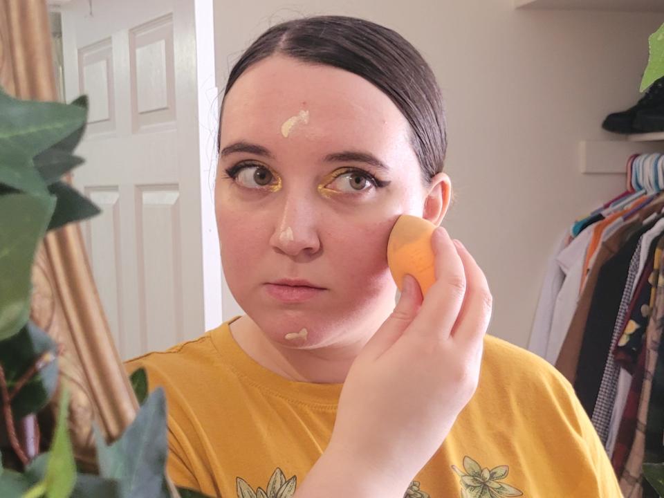 The writer uses an orange beauty sponge to apply foundation to their face