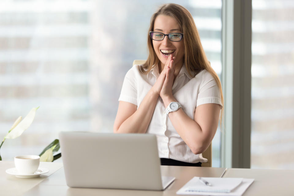 Businesswoman laughing workplace, looking at her laptop screen and clasping her hands together excitedly.