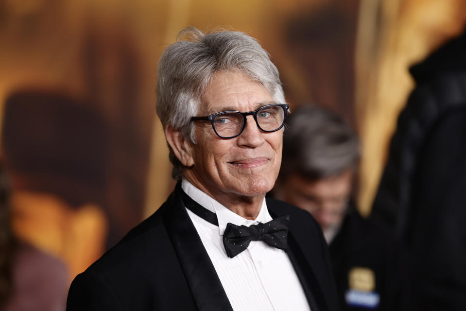 An older man wearing glasses and a black tuxedo with a bow tie on a red carpet, smiling