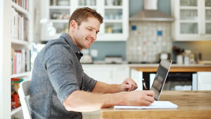 Shot of a young man using a laptop and writing notes at home.