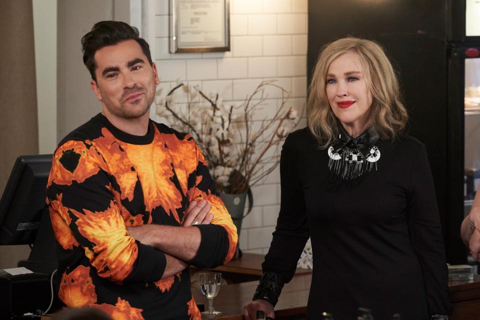 David Rose and Moira Rose from Schitt's Creek stand side by side; he wears a flame-patterned sweater, she in a black dress with a statement necklace