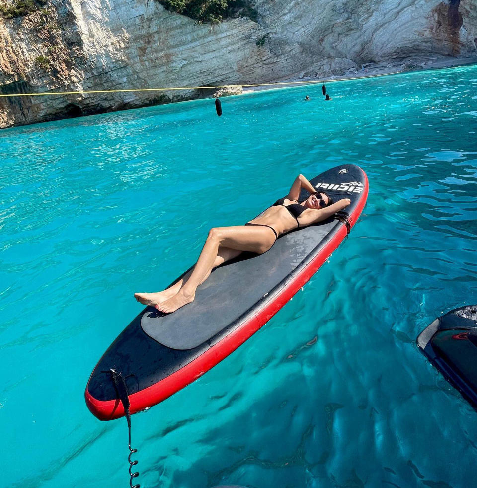 Demi Moore lies on her back on a paddle board in turquoise waters wearing a black bikini