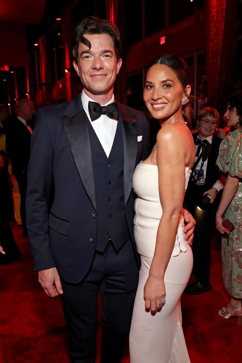 john mulaney and olivia munn pose for a photo in a crowded room, he holds a hand on her waist and wears a navy tuxedo, she wears a white strapless gown