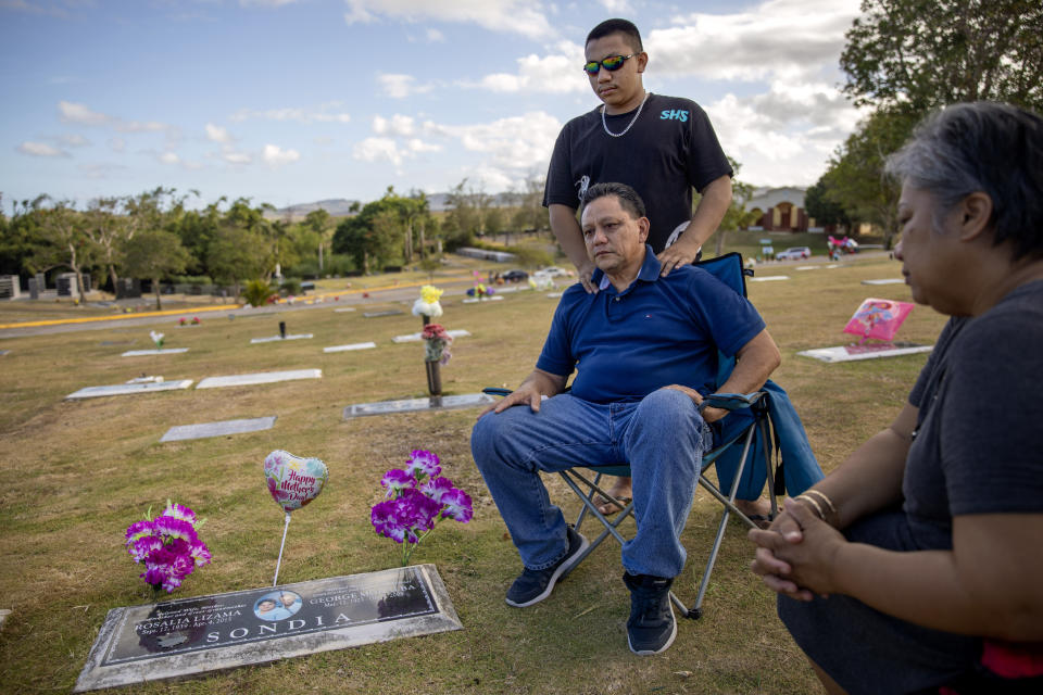 Roland Sondia is comforted by his son, Daniel, 19, while visiting the grave of his mother, Rosalia, on Mother's Day at Our Lady of Peace Memorial Gardens along with his wife, Frances, right, in Yona, Guam, Sunday, May 12, 2019. "She would have been devastated," said Sondia of his mother passing before he made public his story of sexual abuse as a boy by a priest she admired. "I just feel like she needed to know before she left us." (AP Photo/David Goldman)