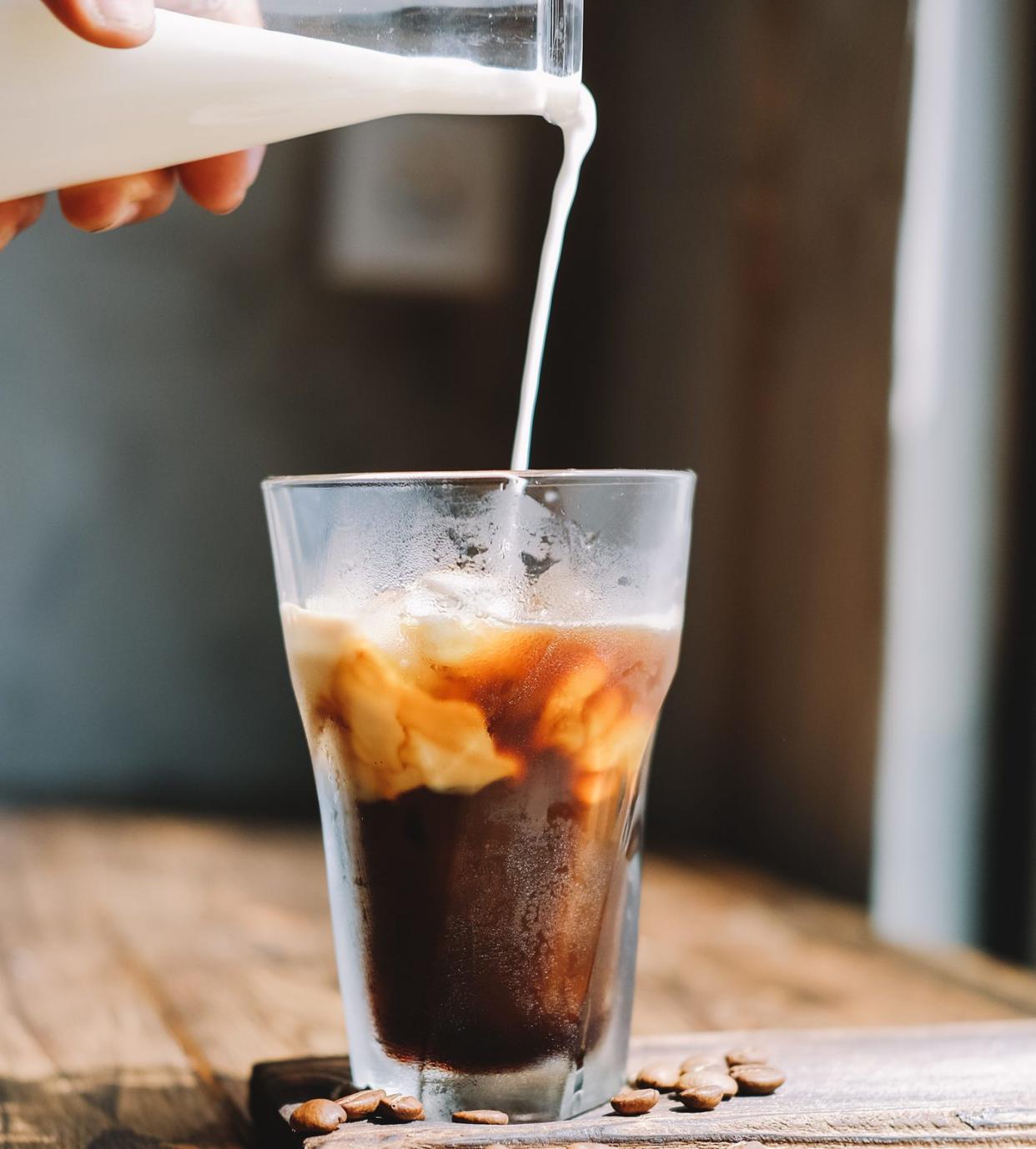 Cropped Hand Pouring Milk In Iced Coffee On Wooden Table