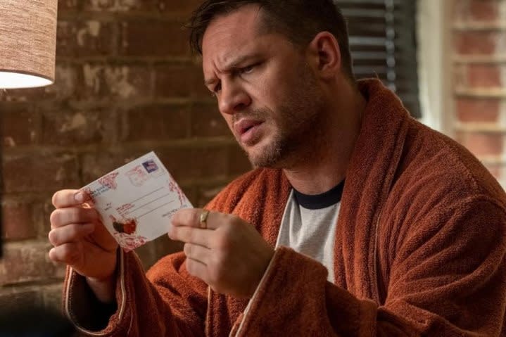 Tom Hardy stares at a bloody postcard while holding it in his hands.