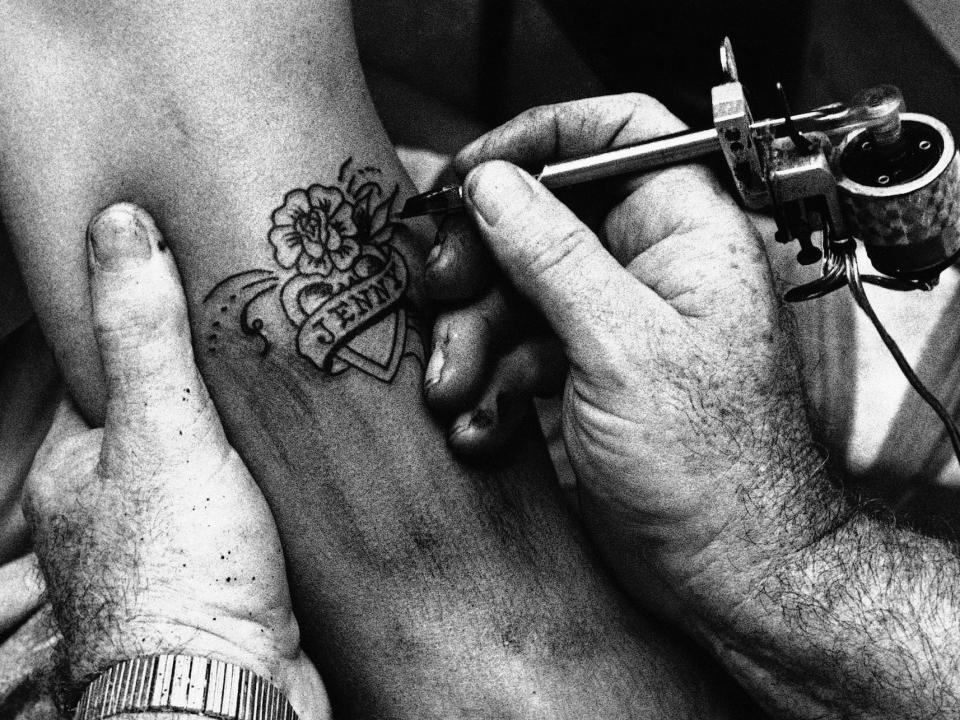 A tattoo artist at a Rhode Island shop etches a design featuring a heart, flowers, and the name "Jenny" on a client's arm.