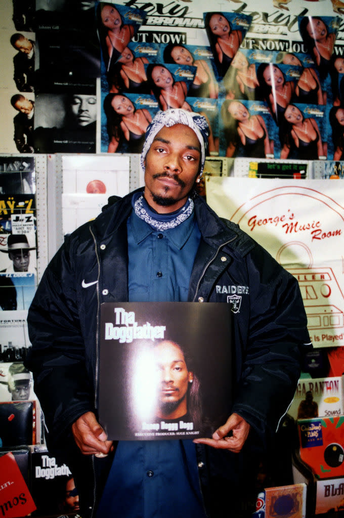 Snoop Dogg holding his album "Tha Doggfather" in a music store with posters in the background