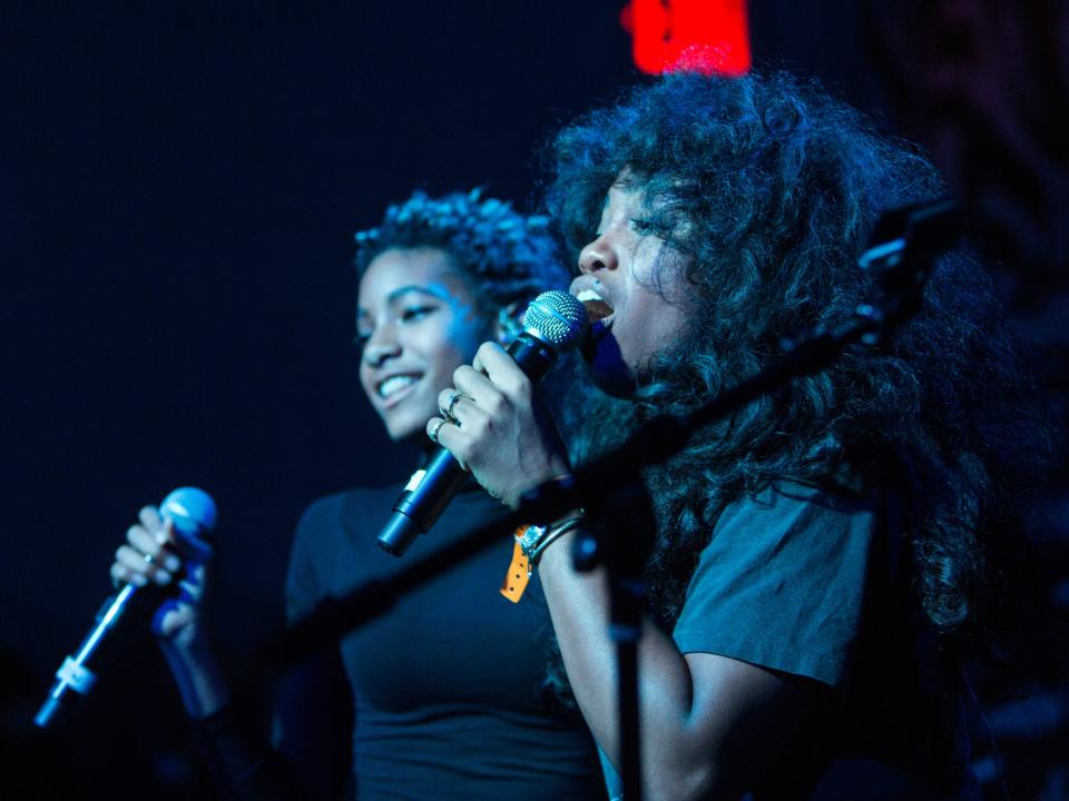 SZA and WILLOW side by side. SZA is closer to the camera. SZA is singing, WILLOW is smiling, facing the crowd, waiting her turn to sing.