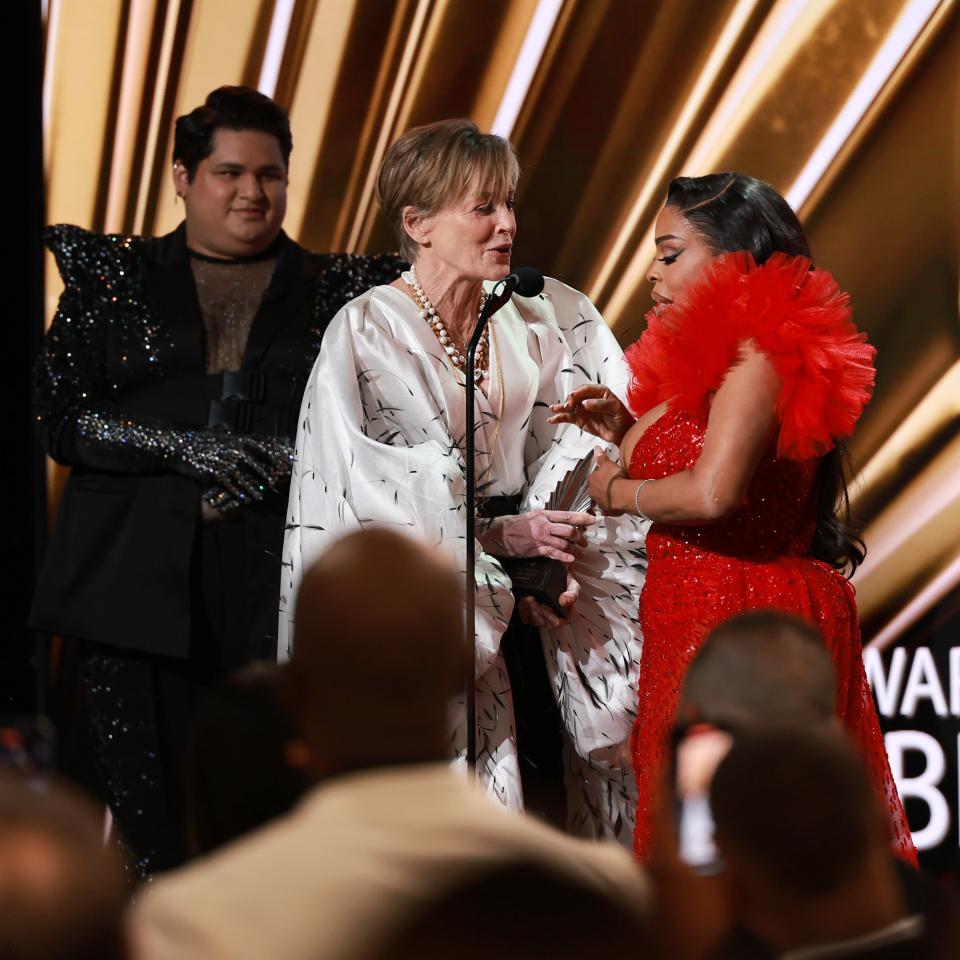 Three people on stage at an award show; one in a ruffled red outfit, another in a patterned blazer, and a presenter speaking