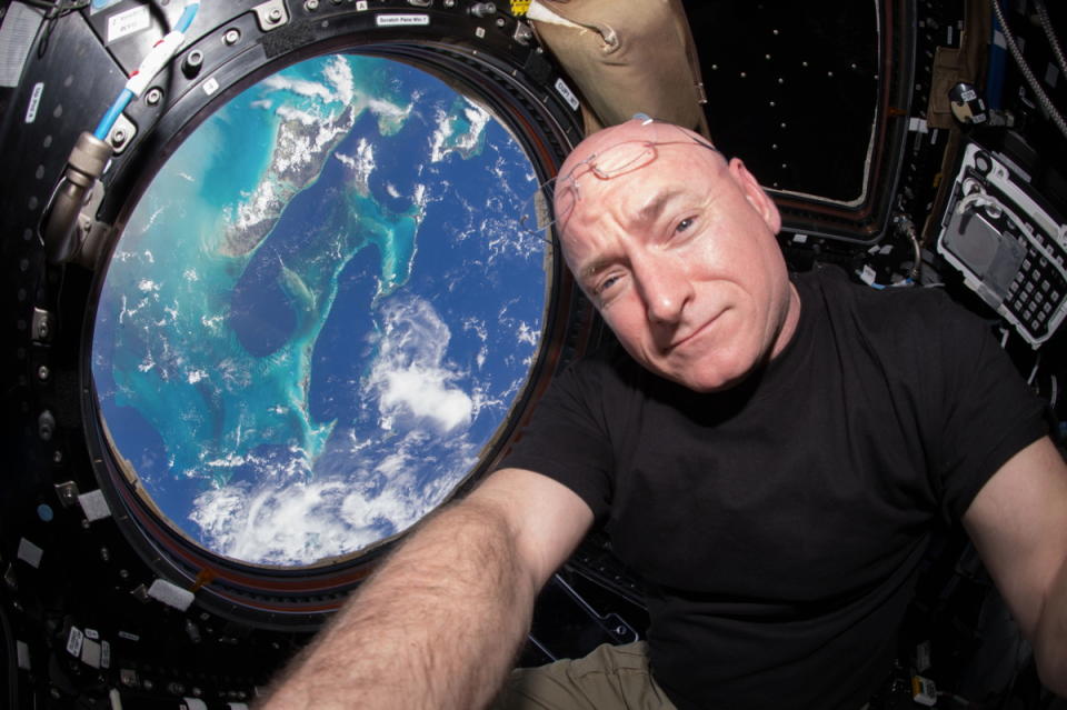a man in a black t-shirt poses in front of a window, through which the earth is visible far below