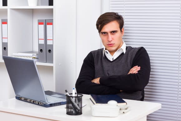 An angry young man with his arms crossed while seated at his desk in front of his laptop.