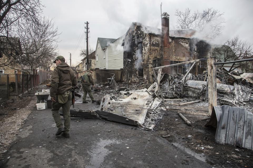 Ukrainian servicemen patrol a bombed-out scene of destruction, with a still-smoking house in the background.