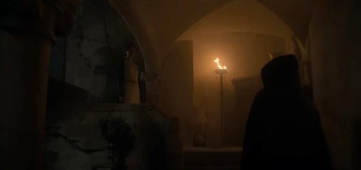 A hooded figure approaches a woman standing on stairs