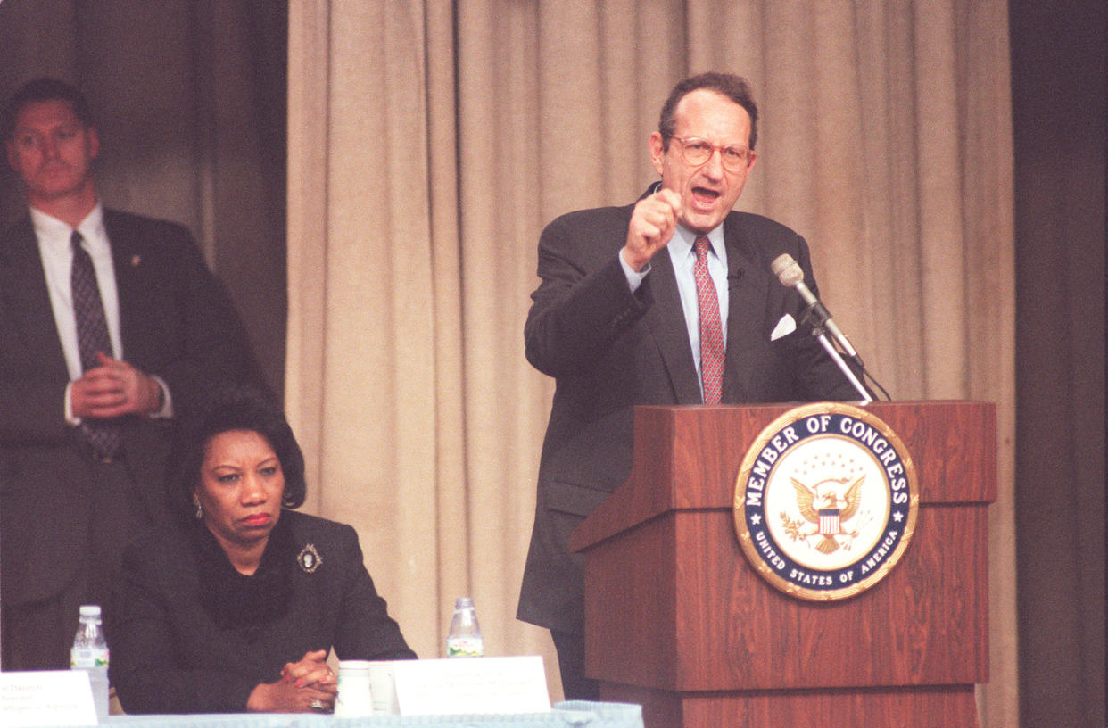 John Deutch, at the podium, with Juanita Millender McDonald on his right, makes an emphatic point at the podium, which is marked: Member of Congress, United States of America.