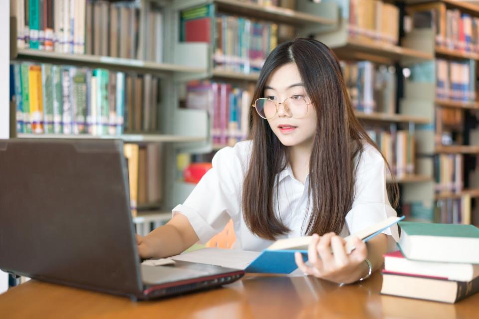 A young woman uses a laptop while holding a book in her hand.