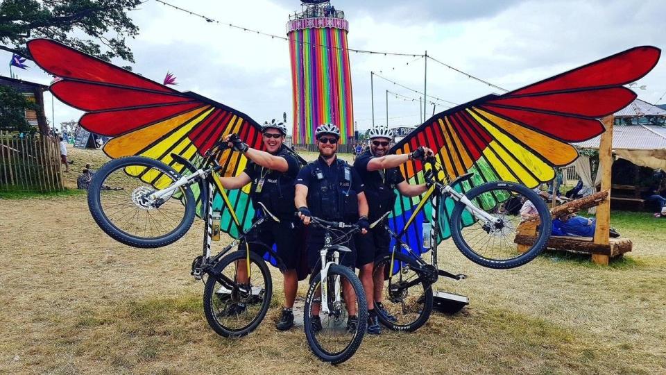 Three policeman and their bikes posing in front of the large colourful wings at Glastonbury