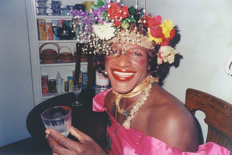 marsha p johnson smiles while sitting in a chair next to a table and cabinet with various household goods, she holds a glass and wears a pink off the shoulder top, necklaces, and a flower crown