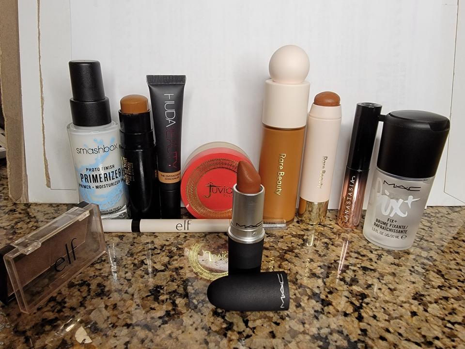 Makeup products arranged on a table, including a Smashbox primer, Huda Beauty concealer, Black Radiance foundation stick, Rare Beauty foundation and bronzer stick, and Mac Fix+