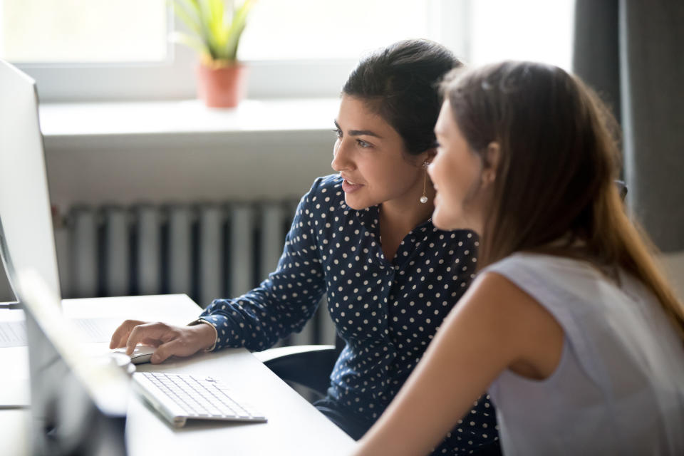 Smiling multiracial millennial girls look at screen work at computer in office together, young diverse female colleagues cooperate using pc, browsing internet or reading news. Collaboration concept