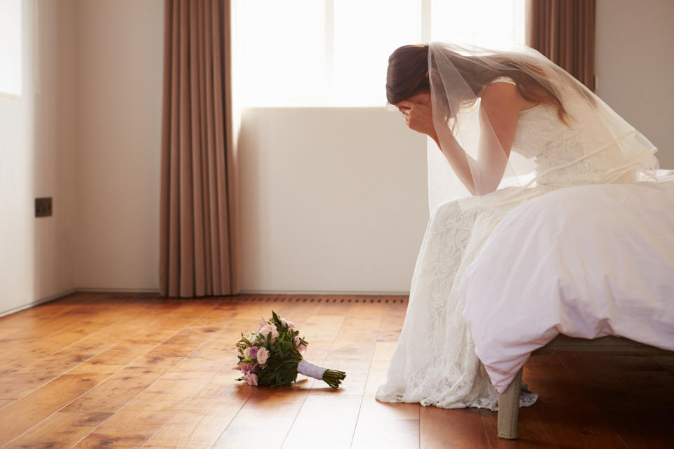 A bride in a white wedding dress and veil sits on a bed, head in hands, looking distressed