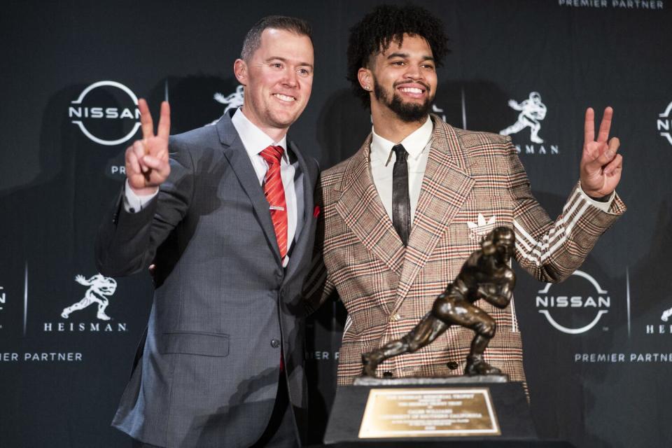 Caleb Williams and Lincoln Riley pose while flashing the USC "victory" symbol while standing next to the Heisman Trophy