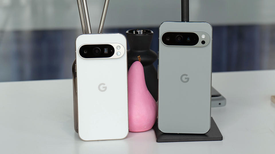 The Pixel 9 Pro and the Pixel 9 Pro XL are propped up on a glass table with a pink pear candle in the background. 