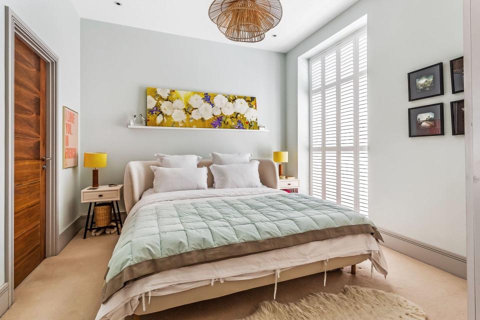 a pale blue bedroom with bed, floral artwork, statement pendant light and a large window with white shutter blinds