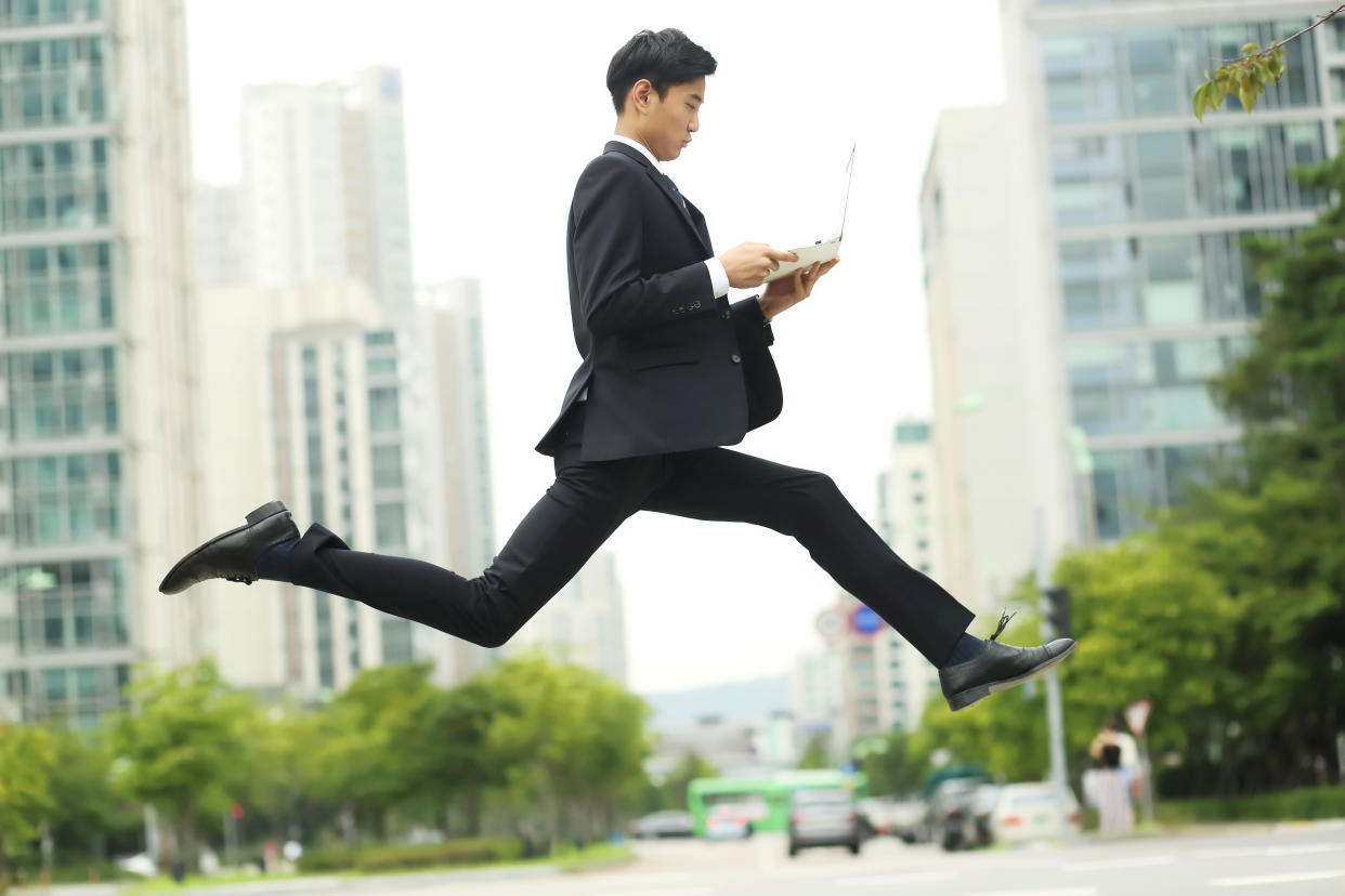 A man in a business suit hopping while holding an opened laptop, over an urban backdrop, illustrating a story on job hopping.