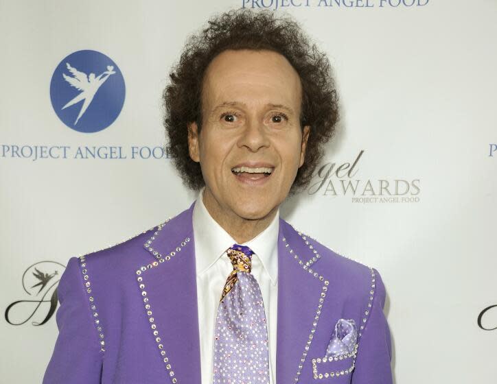 A man with short curly hair in a purple suit with a bedazzled trim and a matching tie smiling with his mouth open