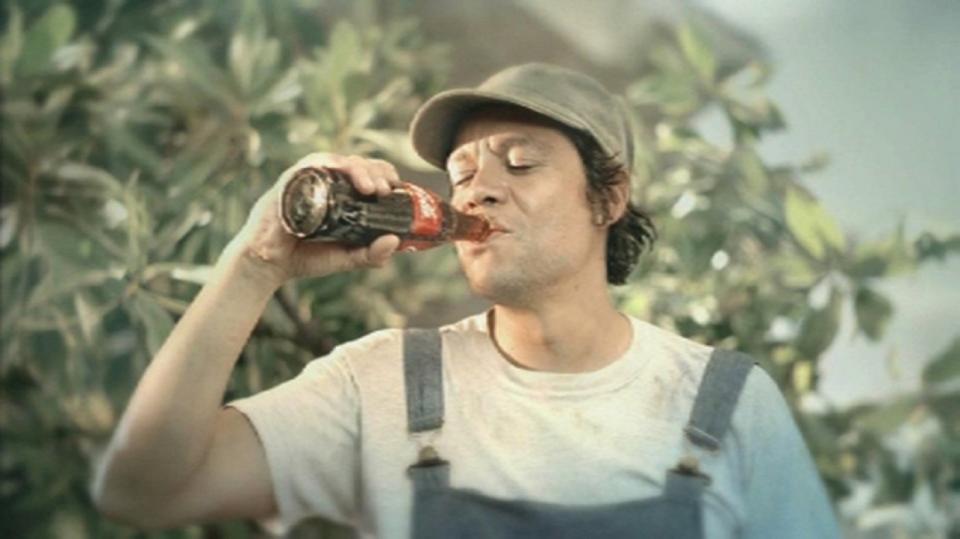 An outdoor worker wearing overalls who's taking a sip from a Coca-Cola bottle.