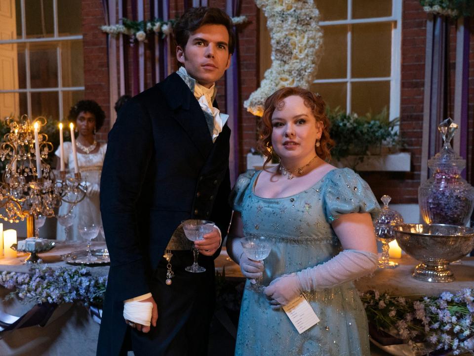 colin bridgerton and penelope featherington standing together at a ball, each holding goblets in their hands and wearing formal clothing