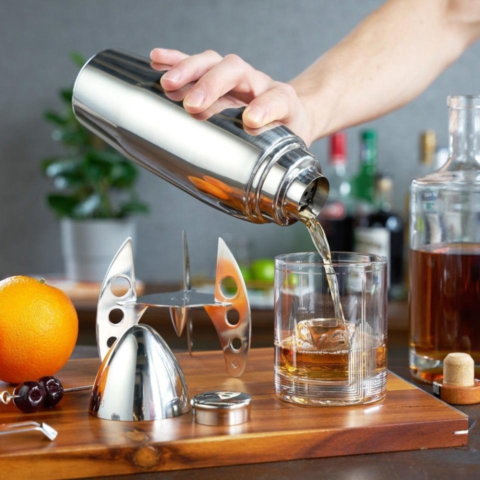 Person pouring liquid from a shaker into a glass, surrounded by cocktail-making tools and ingredients
