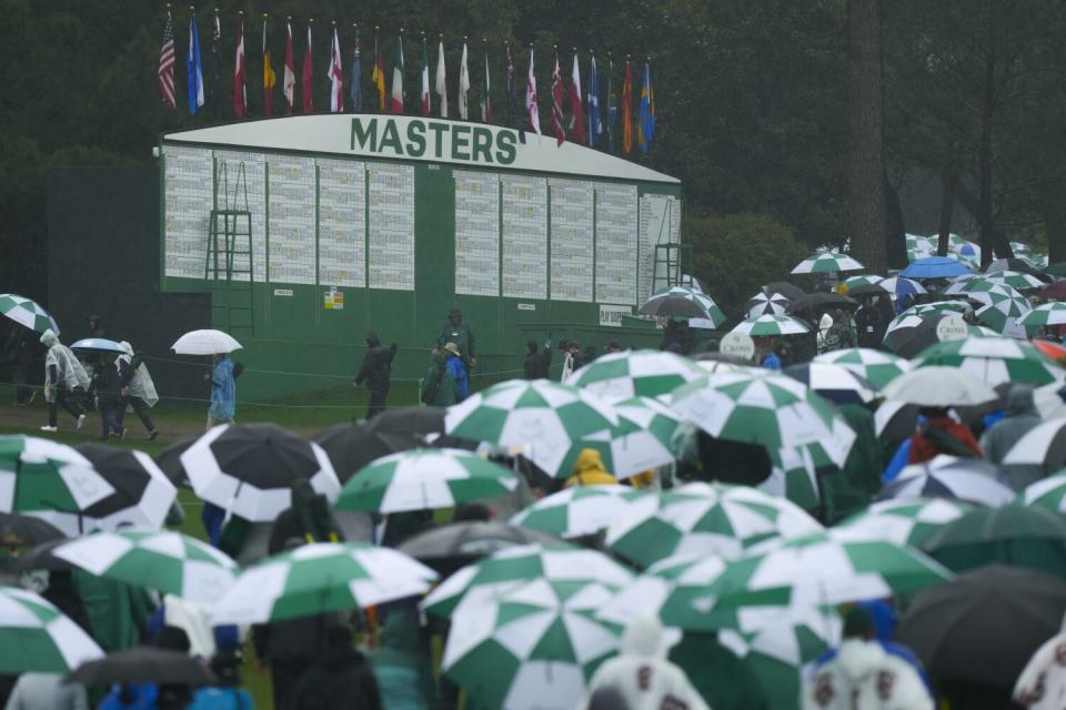Patrons carrying green and white umbrellas walk past the Masters leaderboard.