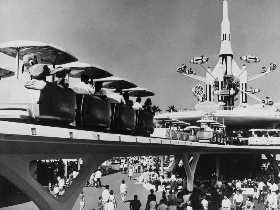 Black and white photo of a monorail in the air with cars on the track and a statue past it with rockets surrounding it.