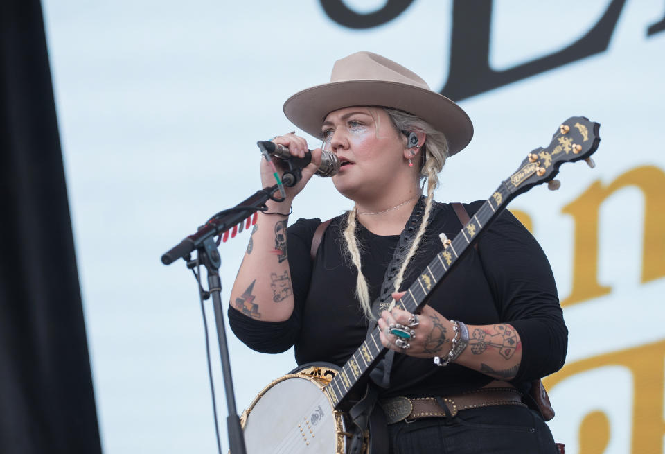 Elle King performs on stage, holding a banjo and singing into a microphone. She wears a wide-brimmed hat, dark shirt, and jeans, with tattoos visible on her arms
